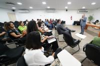 Homem de terno cinza, cabelos grisalhos em pé, falando em frente a homens e mulheres sentados em um auditório