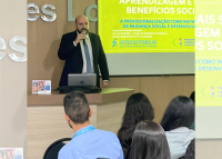 Na imagem, homem calvo, em pé, usando terno preto, gravata cinza e blusa branca, à esquerda dele, um quadro com fundo amarelo e letras brancas a azuis.
