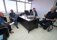 À direita, deputado Paulão, de cabelo grisalho, sentado de camisa preta; à direita dele, atrás de seu birô de trabalho, desembargador Jasiel Ivo, sentada e usando terno cinza, gravata azul-clara e camisa branca; à frente do birô, dois homens morenos sentados.