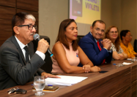 Homem idoso de terno escuro, óculos preto e cabelos grisalhos falando ao microfone, sentado em uma mesa ao lado de uma mulher morena de vestido liso e homem jovem de terno azul e gravata vermelha