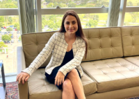 Mulher branca, de cabelos longos, usando terninho branco quadriculado, sentada em um sofá sorrindo
