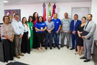 Na imagem, 12 pessoas, sendo seis homens e seis mulheres. Todos então em pé olhando de frente para o ângulo da foto; atrás deles, três bandeiras, sendo a do Brasil a que está no centro, a de Alagoas à direita e a de Maceió à esquerda.