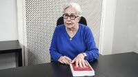 Foto da desembargadora Anne Inojosa. Ela é uma mulher branca de cabelo grisalho, usa óculos pretos e blusa azul. Ela está sentada em uma cadeira preta atrás de um birô preto com a mão esquerda sobre um livro vermelho.