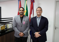 Há dois homens trajados de terno, sendo um com a cor cinza claro e outro escuro. Eles estão de pé posando para uma foto. Em suas costas há três bandeiras, sendo elas, a do Estado de Alagoas, a do Brasil e a do TRT/AL.