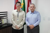 Dois homens de peles claras possam para foto. Eles vestem camisas de mangas longas também claras e calças escuras. As duas pessoas estão à frente de três bandeiras.