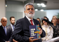 Homem de barba e cabelos brancos vestindo terno cinza e gravata vermelha segura um livro enquanto fala ao microfone