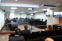 Auditório com mobiliário escuro. Há várias pessoas sentadas, sendo umas em frente a uma bancada e outras na plateia.