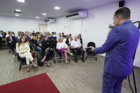 Homem de terno azul, de cabelo castanho escuro,  do lado direito da foto à frente do público proferindo palestra