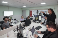 Sala de audiência. Há u,ma homem sentado em frente a um notebook. Ele está digitando. Há quatro pessoas sentadas olhando uma mulher de cabelos longos eescuros utilizando gestual da linguagem de libras.