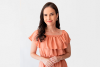 Mulher jovem de pele clara e cabelos longos escuros. Ela veste um vestido de cor clara e tá  posicionada para uma foto.