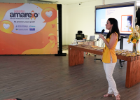 Mulher de cabelo preto, calça branca e blusa amarela com microfone na mão ao lado de um grande banner amarelo com a frase Setembro Amarelo