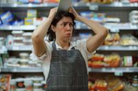 Imagem de uma mulher branca, de cabelo preto, com as duas mãos na cabeça segurando um tablet. A mulher está com cara de constrangimento. Ao fundo, uma prateleira de supermercado cheia de produtos.