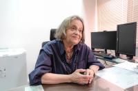 Mulher idoso sentada à mesa; ela tem pelo morena e cabelo grisalho e usa uma blusa azul marinho; ao lado esquerdo dela, a imagem de dois monitores, um em formato horizontal e outro vertical