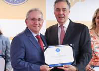 À esquerda, homem branco, de cabelo grisalho, usando paletó azul, gravata cor de vinho; à direita, homem alto, cor branca, usando paletó preto e gravata lilás. Eles estão em pé segurando um diploma branco com bordas pretas
