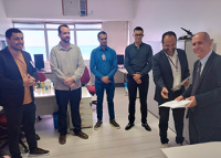 Seis homens em pé; cinco deles estão utilizando calça preta e dois, blazers pretos; à direita, um homem idoso, calvo, magro, de paletó preto e segurando um papel banco.