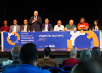 Várias pessoas sentadas. Há uns de ternos e outros sem terno. Há um homem em pé segurando um microfene.