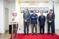 À esquerda, em pé, mulher branca de cabelo grisalho discursando no púlpito com o microfone na mão, à direita, quatro homens em pé usando paletós escuros