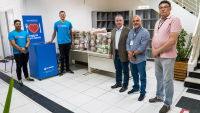 Há cinco homens. Todos estão de pé posan para foto. Há uma caixa coletora e há vários fardos de alimentos em cima de uma mesa.