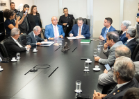 Várias autoridades de paletó sentadas à mesa; em destaque, à esquerda homem magro de cabelo grisalho de paletó preto assinando um termo de cooperação; ao centro da mesa, homem de paletó azul também assinando o documento. 