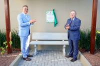 Á esquerda, homem alta de terno azul, ao centro da parede, uma placa de inauguração; à direita, homem baixo, grisalho, de paletó escuro.