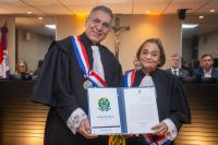 À esquerda, homem alto de cabelo  grisalhos e de toda preta; à direita, mulher morena baixa e idosa também de toga; ambos estão segurando o certificado branco grande.