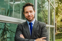 Há um homem branco, com cabelo e barba claros. Ele está vestido com um terno escuro, camisa azul-clara e gravata azul-escura. De braços cruzados, ele sorri.