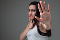 Há uma mulher branca de cabelos longos e escuros. Ela está com o olho roxo e veste uma camiseta branca. A mulher está com uma mão no queixo e outra mão espalmada, como a fazer um gesto de afastamento e de contenção para parara algo.