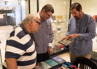 Três homens estão reunidos observando atentamente um porta-retratos. O primeiro veste uma camisa de manga curta com listras azuis e brancas, enquanto os outros dois usam camisas de manga longa em diferentes tons de azul.