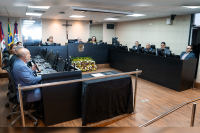 Na foto, vários homens de paletó sentados do lado direito e do lado esquerdo atrás de uma bancada preta, ao centro dessa bancada, um homem de paletó azul e ao lodo esquerdo dele uma mulher de vestido verde e outra mulher de roupa bege.