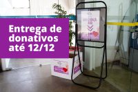 Porta banner preto com cartaz branco com letras na cor lilás. Há uma caixa com a mesma arte do cartaz que está exposto.