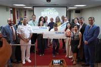 Várias pessoas reunidas para uma foto. Três delas estão segurando um cheque de dimensão bem grande.