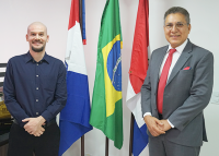 Há dosi homens posando para foto ao lado de três bandeiras. O primeiro homem veste camisa de manga longa e calça escura.. O segundo homem veste terno cinza e usa gravata vermelha.