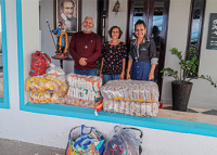 Na imagem, três fardos dos seguintes alimentos: feijão, açúcar e fuba; atrás desse materiais, da esquerda para a direita, um homem de cabelo grisalho e de  blusa longa cor de vinho, uma mulher baixa e morena usando blusa preta florida e outra mulher morena usando saia jeans e blusa cinza de mangas compridas