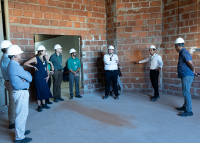Várias pessoas de capcete e botas observam um homem falar. Eles estaõ em uma obra de construção