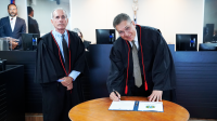 Há um homem de toga em pé, do lador esquerdo. Ao seu lado há outro homem inclinado, também trajando toga. Ele assina um papel.