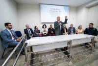 Homens e mulheres sentados atrás de uma mesa e um homem de terno cinza falando à frente