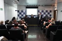 Homem de terno preto e camisa branca em pé do lado direito do auditório falando para várias pessoas que estão sentadas de frente para ele.