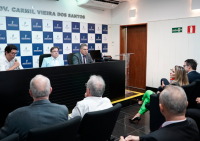 Há três homens sentados. Eles estão de frente a uma plateia que asssite um dos homens falando.