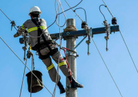 Foto de um eletricista de costa, de macacão cinza e capacete branco, em cima de um poste