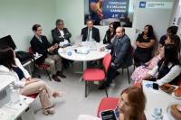 Vá rias pessoas reunidas em uma sala. Há uma mesa rendonda no centro da sala.