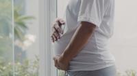 Perfil de mulher gestante segurando a barriga. Ela veste blusa branca e calça cinza.