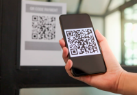 Há uma mão de pessoa branca com o celular na mão com um QR Code na tela.