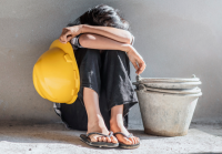 Criança sentada no chão com as costas apoiadas em uma parede. Ela usa calça preta e está com as duas pernas dobradas e os dois braços por cima dos joelhos cobrindo o rosto e segurando um capacete amarelo com a mão esquerda