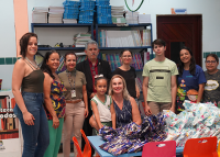 Várias pessoas reunidas para uma foto. Há embalagens em cima das mesas escolares infantis.