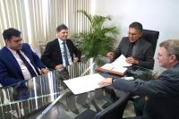 A imagem mostra quatro homens sentados ao redor de uma mesa de vidro em uma reunião. Eles estão vestidos formalmente, usando ternos e gravatas. O homem ao centro, que parece estar conduzindo a reunião, veste um terno escuro e óculos, segurando um papel em suas mãos. À sua direita, um homem de terno azul e gravata listrada tem uma expressão séria, enquanto outro homem ao lado dele, de terno preto e gravata listrada, sorri levemente. 