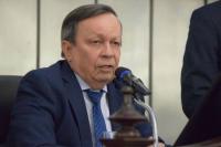 Homem branco de terno e gravata azul escura. Ele está sentado e tem a sua frente um microfone.
