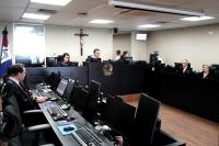 Há várias pessoas reunidas em volta de uma bancada. Todos vestem togas pretas. Eles têm, à sua frente, microfones e notebooks.
