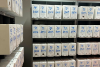 A imagem mostra um arquivo físico organizado em estantes de metal cinza, contendo diversas caixas brancas de papelão identificadas com numeração e inscrições em azul. Cada caixa tem um espaço para anotações na parte frontal e está disposta de maneira ordenada nas prateleiras. A inscrição "56 VT" aparece repetidamente nas caixas, seguida de números sequenciais, como "1865", "1866", "1867" e assim por diante. O ambiente sugere um arquivo institucional, possivelmente de documentos jurídicos ou administrativos
