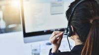 Imagem da cabeça de uma mulher branca de frente ao computador com um fone de ouvido; ela tem cabelos pretos e na imagem só mostra a parte de trás da cabeça dela.