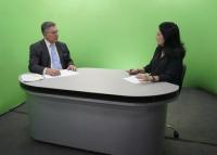 Sentados à mesa da entrevista, homem moreno de cabelos grisalhos, usando óxculos, de paletó preto, gravata azul-clara; à direita, mulher branca de cabelos pretos e blusa preta